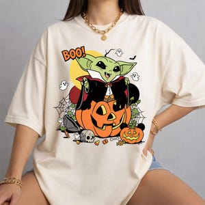 May include: A cream-colored t-shirt featuring a graphic of a green creature dressed as a vampire sitting on a large orange jack-o'-lantern. The design includes the text BOO!, small ghosts, a spider web, a skull, and a smaller pumpkin with mouse ears.