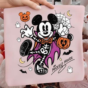 Puede incluir: Una camiseta rosa claro con un gráfico de Mickey Mouse disfrazado de vampiro, con estampado de esqueleto, capa morada y un cubo de calabaza naranja. El diseño incluye pequeños fantasmas, murciélagos, una telaraña y el texto Mickey Mouse.
