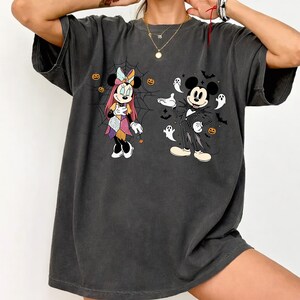 Peut inclure: Un t-shirt gris anthracite à manches courtes avec une impression graphique de Minnie Mouse vêtue d'une robe en patchwork dans une toile d'araignée et de Mickey Mouse en costume rayé entouré de fantômes blancs, de chauves-souris et de citrouilles.