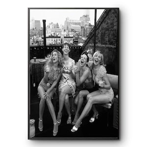 May include: A black and white photograph featuring four individuals sitting together on a rooftop terrace, laughing while holding glasses. The background displays a cityscape with various buildings under a bright sky, framed by a dark border.