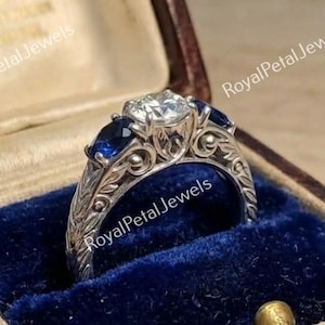 May include: A silver ring featuring a central round white gemstone flanked by two smaller blue oval gemstones. The band displays intricate engraved scrollwork patterns along the sides and sits within a dark blue velvet-lined jewelry box.