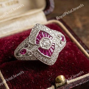May include: A silver-toned ring featuring a central round diamond surrounded by baguette-cut red rubies and small pavé-set diamonds in a geometric arrangement. The ring is displayed inside a dark burgundy velvet-lined jewelry box.