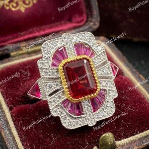May include: A vintage-style ring with a large, rectangular, deep red gemstone at the center, surrounded by smaller pink and clear stones. The ring has a gold setting around the central stone and a silver band.