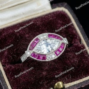 Puede incluir: Un anillo de compromiso con corte marquesa con una piedra central transparente, rodeada de piedras rojas cuadradas y pequeñas piedras transparentes. El anillo está engastado en una banda plateada y descansa en una caja de terciopelo burdeos oscuro.