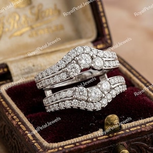 Op de afbeelding: Twee witgouden diamanten ringen, gepresenteerd in een bordeauxrode fluwelen sieradendoos. De ringen hebben meerdere rijen kleine diamanten en grotere centrale stenen. De doos heeft gouden accenten en de tekst "Royal Petal Jewels".