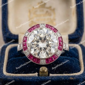 May include: A ring featuring a large, round, clear center stone surrounded by a halo of alternating square-cut red stones and small, round, clear accent stones. The metal band is silver-toned, and the ring is set within a dark blue velvet box.