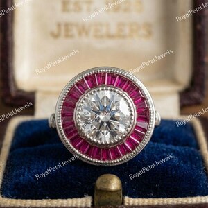 May include: A silver ring with a large, round, clear gemstone at the center, surrounded by a ring of smaller, rectangular, red gemstones. The ring is set in a vintage-style jewelry box with a blue velvet interior. The text "Royal Petal Jewels" is visible.