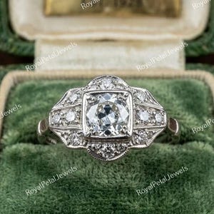Pode incluir: Um anel de tom prateado com uma pedra central redonda de corte brilhante cravada num quadrado, rodeada por uma moldura decorativa de pedras redondas mais pequenas. A banda de metal tem detalhes intrincados e bordas milgrain.