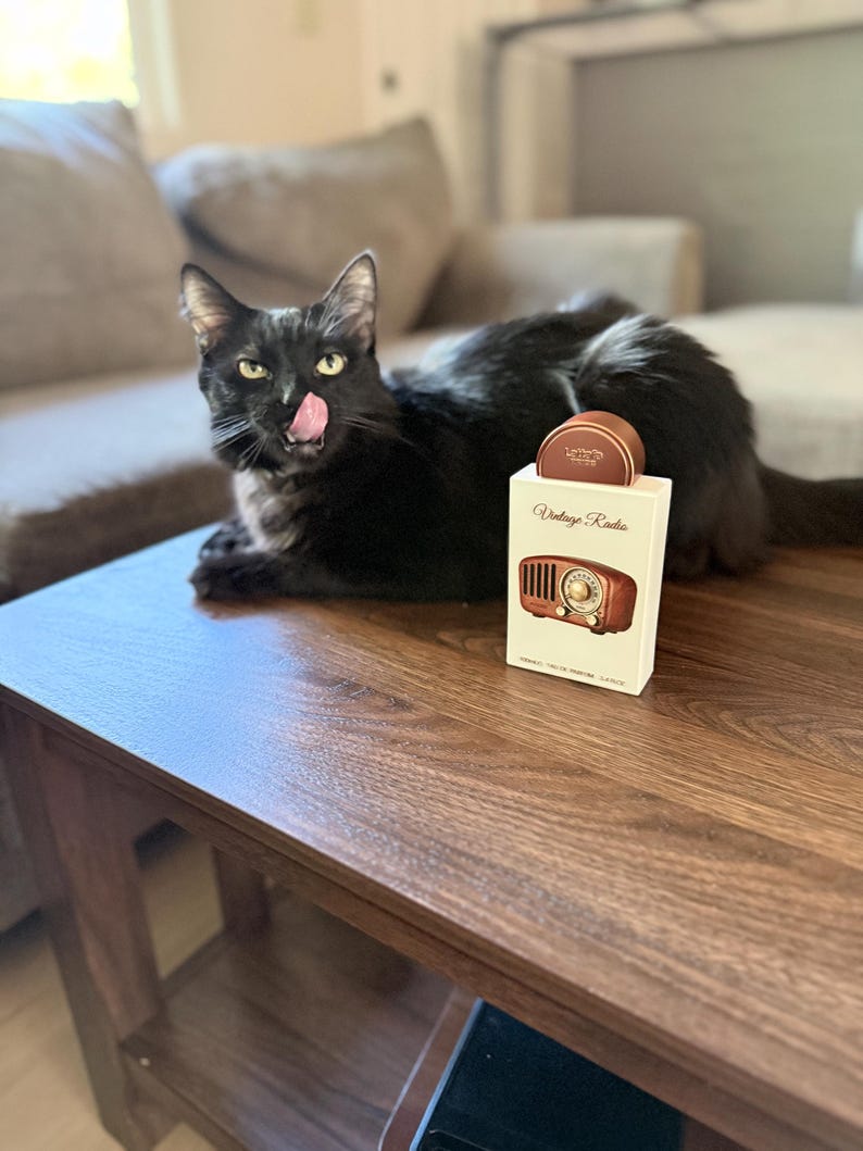 May include: A black cat with its tongue out rests on a wooden table next to a box of coasters. The box is white with the words "Vintage Radio" and a picture of a vintage radio. The coasters are brown.
