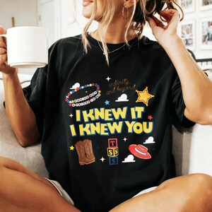Puede incluir: Una camiseta negra de manga corta con un estampado gr&aacute;fico colorido que incluye el texto "I KNEW IT I KNEW YOU" en amarillo, junto a ilustraciones de una pulsera de cuentas, una estrella de sheriff, botas vaqueras, bloques y un sombrero rojo.