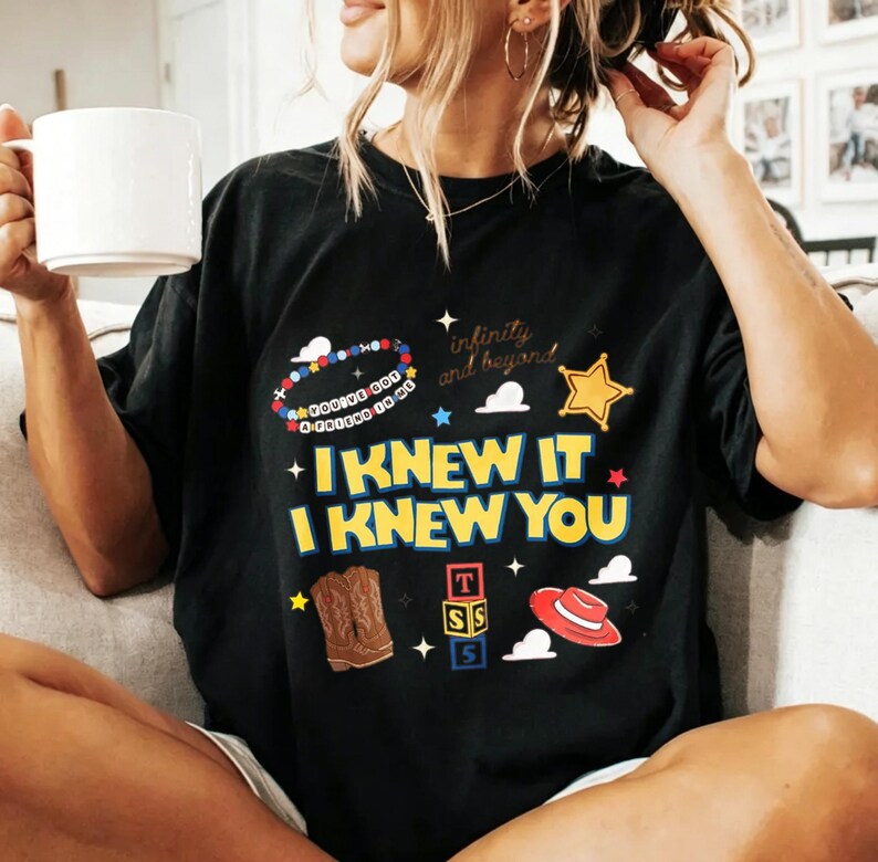 Puede incluir: Una camiseta negra de manga corta con un estampado gr&aacute;fico colorido que incluye el texto "I KNEW IT I KNEW YOU" en amarillo, junto a ilustraciones de una pulsera de cuentas, una estrella de sheriff, botas vaqueras, bloques y un sombrero rojo.