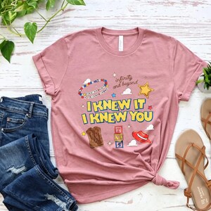 Puede incluir: Una camiseta de manga corta de color malva con el texto I KNEW IT I KNEW YOU en letras amarillas y azules. El gr&aacute;fico incluye una pulsera de cuentas, una estrella amarilla, botas vaqueras marrones, bloques de letras y un sombrero rojo con el texto infinity and beyond.