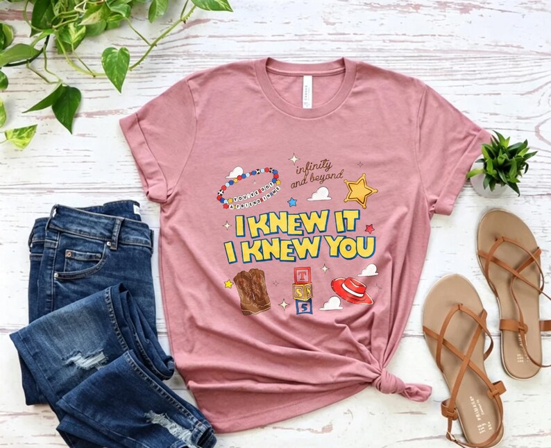 Puede incluir: Una camiseta de manga corta de color malva con el texto I KNEW IT I KNEW YOU en letras amarillas y azules. El gr&aacute;fico incluye una pulsera de cuentas, una estrella amarilla, botas vaqueras marrones, bloques de letras y un sombrero rojo con el texto infinity and beyond.