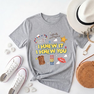 Puede incluir: Una camiseta gris jaspeado de manga corta con un estampado gr&aacute;fico que incluye el texto "I KNEW IT I KNEW YOU" en amarillo, "infinity and beyond" en marr&oacute;n y "YOU'VE GOT A FRIEND IN ME" en una pulsera de cuentas, junto a botas, un sombrero y bloques.