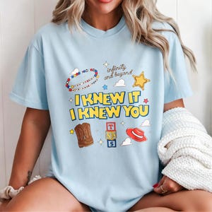 Puede incluir: Una camiseta azul claro con un estampado gr&aacute;fico que incluye el texto "I KNEW IT I KNEW YOU" y "infinity and beyond". El dise&ntilde;o muestra una pulsera de cuentas, una estrella amarilla, botas vaqueras marrones, bloques y un sombrero rojo.