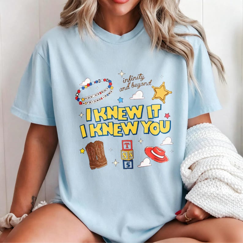 Puede incluir: Una camiseta azul claro con un estampado gr&aacute;fico que incluye el texto "I KNEW IT I KNEW YOU" y "infinity and beyond". El dise&ntilde;o muestra una pulsera de cuentas, una estrella amarilla, botas vaqueras marrones, bloques y un sombrero rojo.