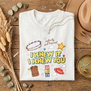 Puede incluir: Una camiseta blanca con un estampado gr&aacute;fico colorido con el texto "I KNEW IT I KNEW YOU", "infinity and beyond" y "YOU'VE GOT A FRIEND IN ME". El dise&ntilde;o incluye ilustraciones de una pulsera de cuentas, una estrella, botas vaqueras, bloques y un sombrero.