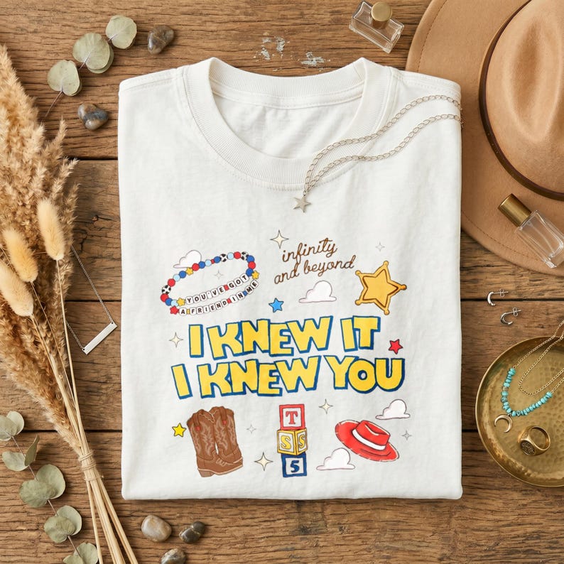 Puede incluir: Una camiseta blanca con un estampado gr&aacute;fico colorido con el texto "I KNEW IT I KNEW YOU", "infinity and beyond" y "YOU'VE GOT A FRIEND IN ME". El dise&ntilde;o incluye ilustraciones de una pulsera de cuentas, una estrella, botas vaqueras, bloques y un sombrero.