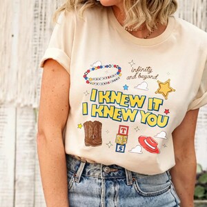 Puede incluir: Una camiseta de color crema con un estampado gr&aacute;fico que incluye el texto "I KNEW IT I KNEW YOU" y "infinity and beyond". El dise&ntilde;o presenta una pulsera de cuentas, una estrella amarilla, botas vaqueras marrones, bloques y un sombrero rojo.
