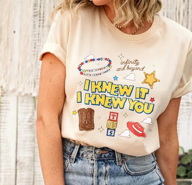 Puede incluir: Una camiseta de color crema con un estampado gr&aacute;fico que incluye el texto "I KNEW IT I KNEW YOU" y "infinity and beyond". El dise&ntilde;o presenta una pulsera de cuentas, una estrella amarilla, botas vaqueras marrones, bloques y un sombrero rojo.