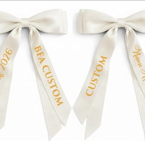 May include: Two ivory satin bows with long, angled tails. One bow has "Class of 2026" and "BFA CUSTOM" printed in gold. The other bow has "CUSTOM" and "Name Here" printed in gold. Graduation and custom event decor.