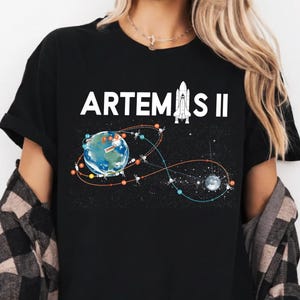 May include: Black t-shirt featuring the text "ARTEMIS II" above a graphic of Earth, a rocket, and a celestial design. The shirt has a crew neck and short sleeves. The word "Black" is in a black oval at the bottom of the shirt.