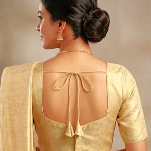 Könnte beinhalten: Eine goldfarbene Bluse mit quadratischem Ausschnitt und kurzen Ärmeln. Der Rücken hat einen Bindverschluss mit Quasten. Die Frau trägt goldene Ohrringe und eine Halskette. Das Haar ist zu einem Dutt frisiert.