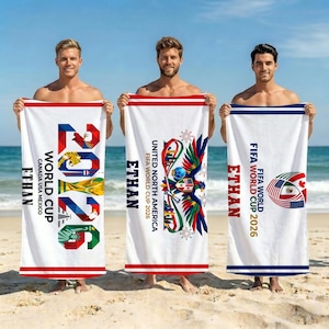 Könnte beinhalten: Drei weiße, rechteckige Strandtücher mit Motiven zur FIFA Fußball-Weltmeisterschaft 2026. Das erste Handtuch zeigt die Zahl 2026 mit bunten Grafiken, den Namen ETHAN und Text. Das zweite zeigt ein Adler-Emblem und Text. Das dritte zeigt eine Fußballgrafik mit Flaggen und Text.