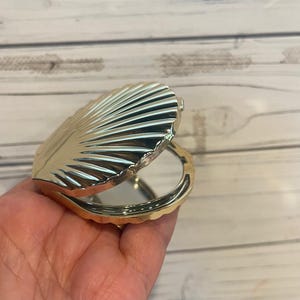 May include: A gold-colored, shell-shaped compact mirror. The compact is open, revealing a circular mirror. The shell design features detailed, ridged lines radiating from the center. The compact is held in a hand against a light wood background.