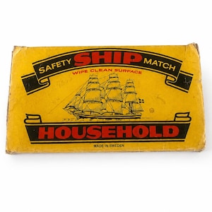 May include: A rectangular yellow matchbox featuring a black and red graphic of a sailing ship. The text reads SAFETY SHIP MATCH, WIPE CLEAN SURFACE, HOUSEHOLD, and MADE IN SWEDEN. The box has a worn, vintage appearance with visible creases and edges.