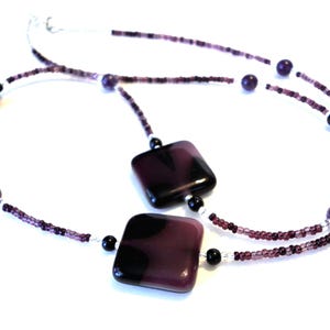 May include: A necklace featuring a double strand design with small purple beads and larger purple and black square and round beads. The necklace has a silver-colored clasp.