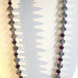 Puede incluir: Un collar largo hecho de cuentas de vidrio facetadas en tonos de morado, verde y gris. Las cuentas est&aacute;n enhebradas en un hilo transparente y el collar se muestra en un maniqu&iacute; blanco.