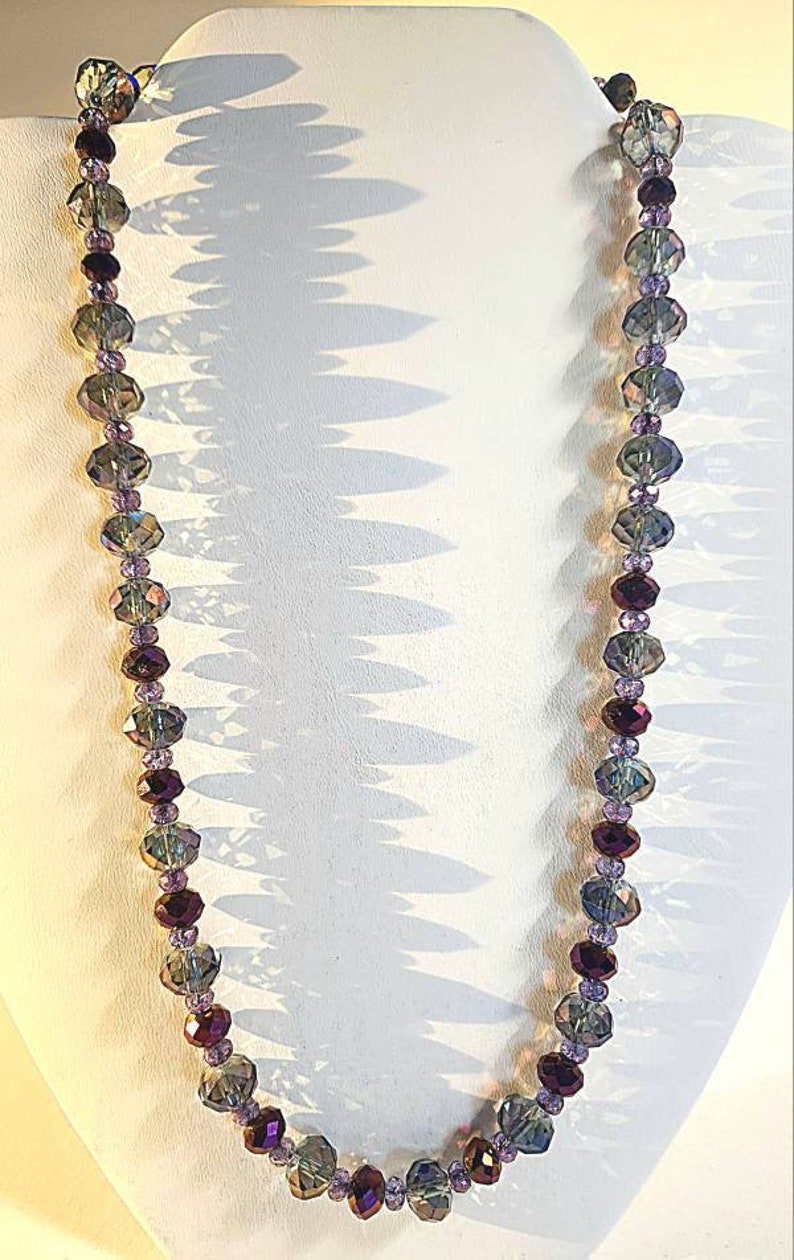 Puede incluir: Un collar largo hecho de cuentas de vidrio facetadas en tonos de morado, verde y gris. Las cuentas est&aacute;n enhebradas en un hilo transparente y el collar se muestra en un maniqu&iacute; blanco.