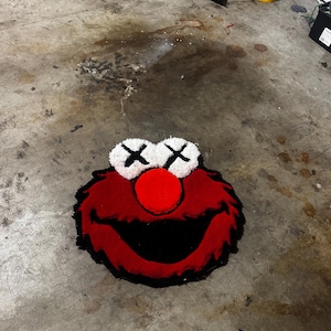 May include: A tufted rug shaped like a character head with red fur, a large red nose, and a wide black mouth. The eyes are white circles with black X markings. The rug features a textured, plush surface and a distinct outline, capturing the iconic facial features of the character in a bold, graphic style.