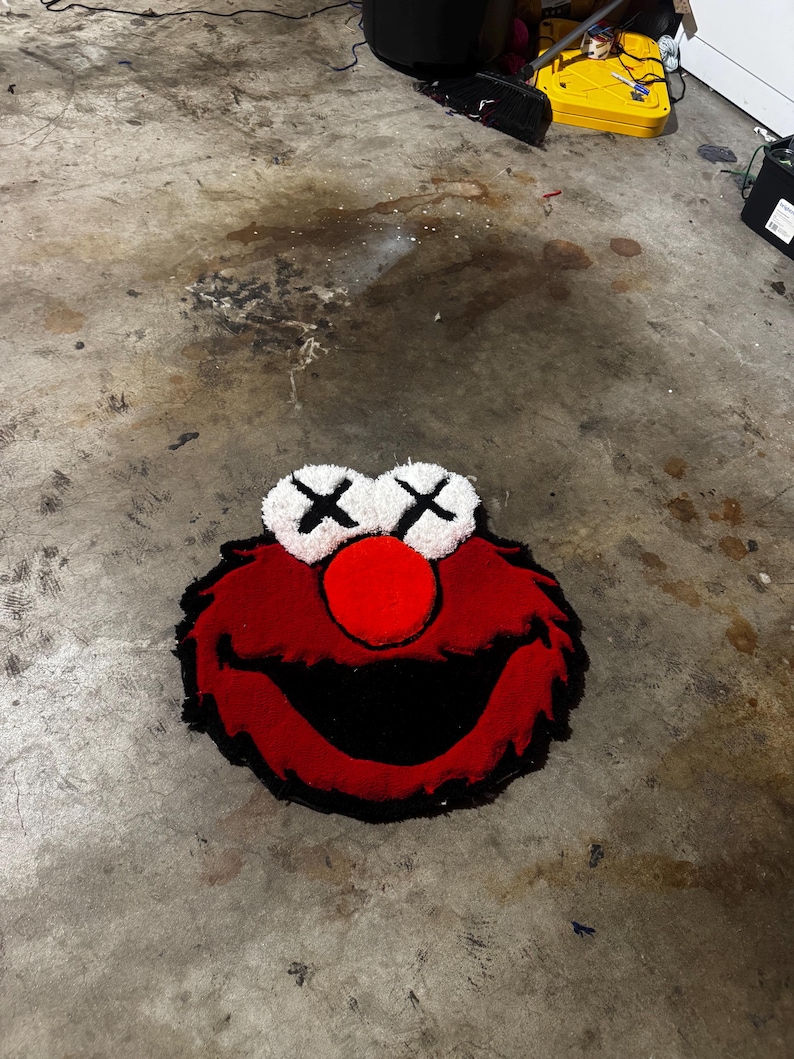 May include: A tufted rug shaped like a character head with red fur, a large red nose, and a wide black mouth. The eyes are white circles with black X markings. The rug features a textured, plush surface and a distinct outline, capturing the iconic facial features of the character in a bold, graphic style.