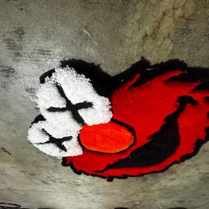 May include: A tufted rug featuring a stylized character face with bright red fur, a large orange oval nose, and two white eye patches marked with black X symbols. The design is outlined in black, creating a bold, high-contrast graphic appearance.