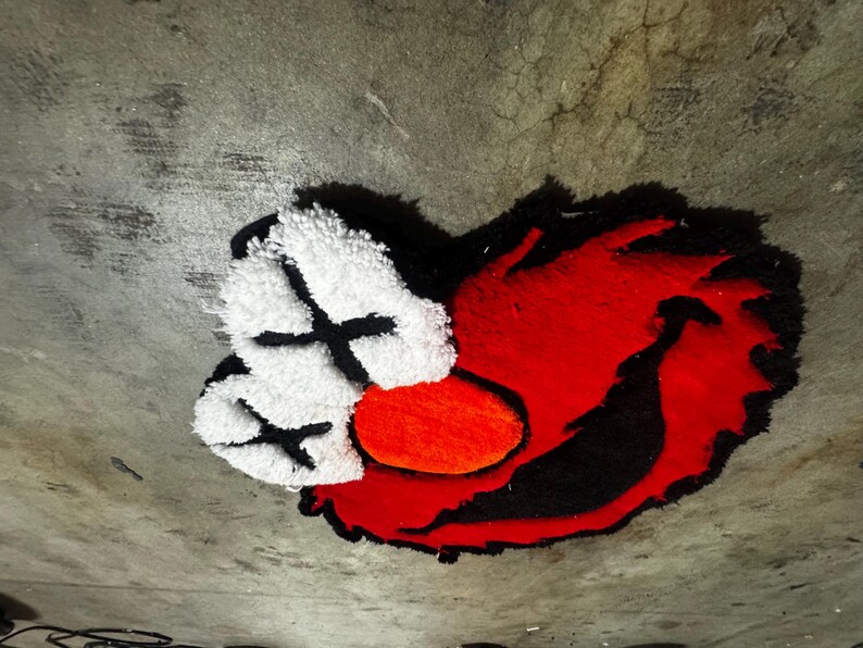 May include: A tufted rug featuring a stylized character face with bright red fur, a large orange oval nose, and two white eye patches marked with black X symbols. The design is outlined in black, creating a bold, high-contrast graphic appearance.