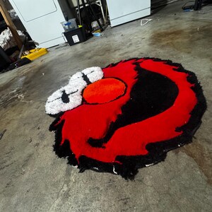 May include: A tufted rug featuring a stylized character face with a large red circular nose, two white eyes with black cross-shaped pupils, and a thick black outline surrounding the vibrant red facial features, all crafted with a textured pile.
