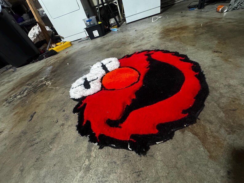 May include: A tufted rug featuring a stylized character face with a large red circular nose, two white eyes with black cross-shaped pupils, and a thick black outline surrounding the vibrant red facial features, all crafted with a textured pile.