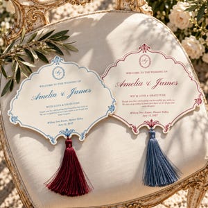 Puede incluir: Dos abanicos de boda con bordes festoneados y cenefas ornamentadas. Uno tiene texto azul y una borla roja oscura, el otro texto rojo y una borla azul. Ambos muestran el texto: Welcome to the wedding of Amelia & James, Willow Tree Estate, June 12, 2027.