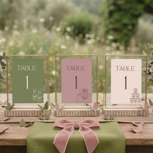 May include: Three rectangular table number signs in gold metal frames with ribbed wooden bases. The left sign is sage green with two champagne flutes, the centre is dusty rose with a table illustration, and the right is cream with a champagne tower.