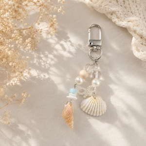 May include: A silver keychain with a decorative charm featuring a seashell, pearls, and beads. The charm includes a spiral seashell, a white scallop shell, and various beads in white, cream, and blue. The keychain is designed with a beach or coastal theme.