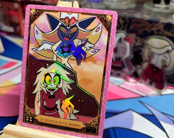 May include: A rectangular trading card with a pink border featuring two animated characters, one with yellow hair and a red outfit, and another with a tall hat and wings. The card includes the text IT STARTS WITH SORRY and is displayed on a small wooden easel.