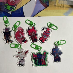 May include: A collection of eight acrylic keychains featuring stylized cartoon characters with button eyes, each attached to a bright green plastic lobster clasp. The characters are depicted in various outfits and colors, including red, white, and black.