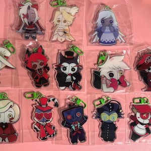 May include: A collection of thirteen individual acrylic character keychains, each enclosed in a clear plastic sleeve with a green plastic lobster clasp. The characters feature button eyes and stylized outfits in various colors including red, black, white, and blue.