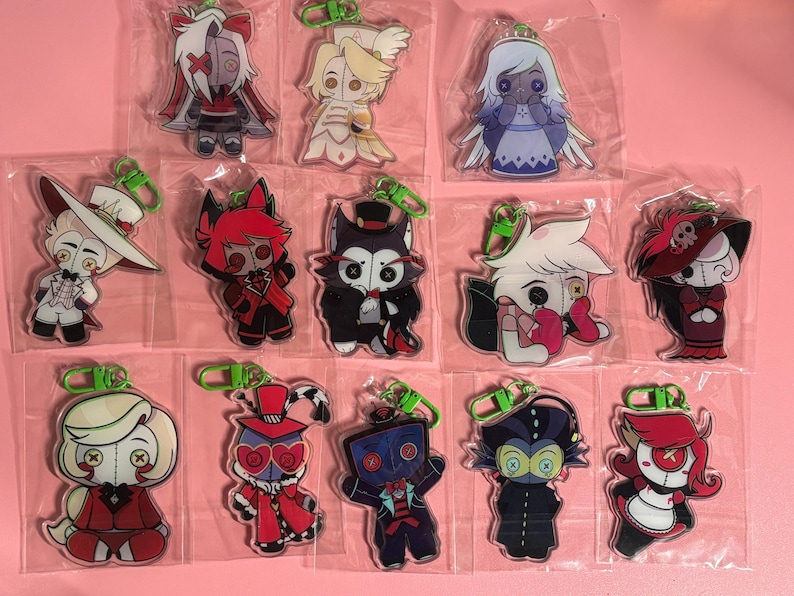May include: A collection of thirteen individual acrylic character keychains, each enclosed in a clear plastic sleeve with a green plastic lobster clasp. The characters feature button eyes and stylized outfits in various colors including red, black, white, and blue.