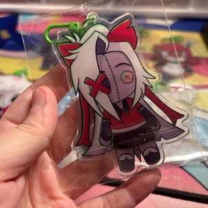 May include: A clear acrylic keychain featuring a stylized character with white hair, a red X over one eye, and a green button over the other. The character wears a red shirt, dark skirt, and has a green plastic lobster clasp attached at the top.