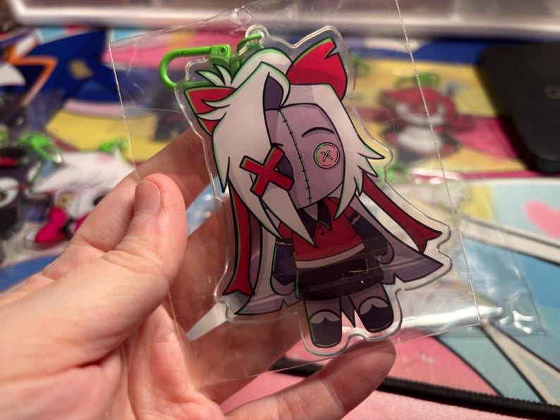 May include: A clear acrylic keychain featuring a stylized character with white hair, a red X over one eye, and a green button over the other. The character wears a red shirt, dark skirt, and has a green plastic lobster clasp attached at the top.