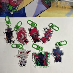May include: A collection of seven clear acrylic keychains featuring various cartoon character designs with vibrant pink, red, black, and white color schemes. Each keychain is attached to a bright green plastic lobster clasp hook for easy attachment.