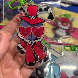 May include: A clear acrylic keychain featuring a cartoon character with a purple head, heart-shaped glasses with button-like lenses, and a red top hat. The character wears a red coat with white fur trim and has a green plastic lobster clasp attached.